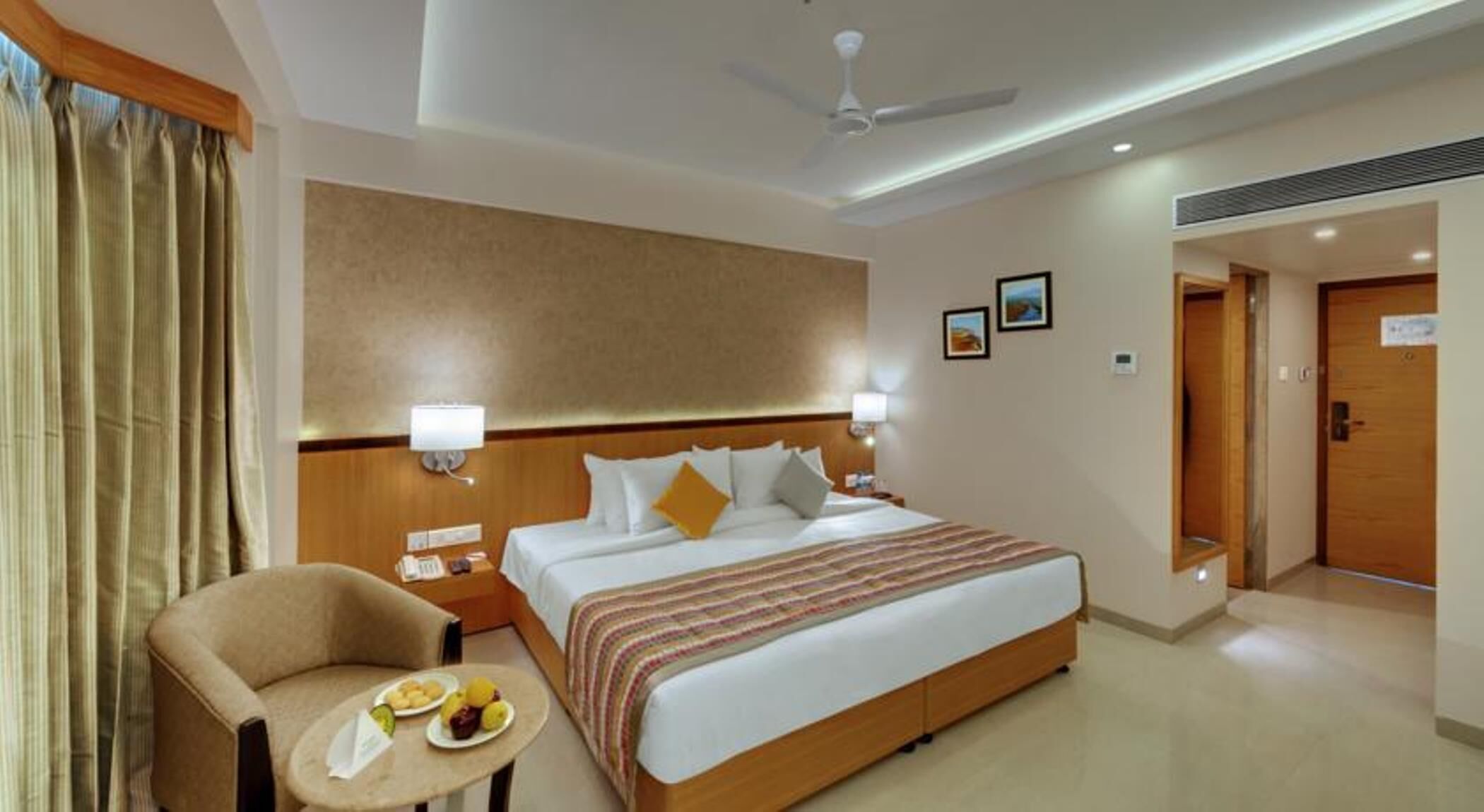 Une chambre d’hôtel avec un lit, des lampes de chevet, une chaise avec une petite table et un ventilateur de plafond.