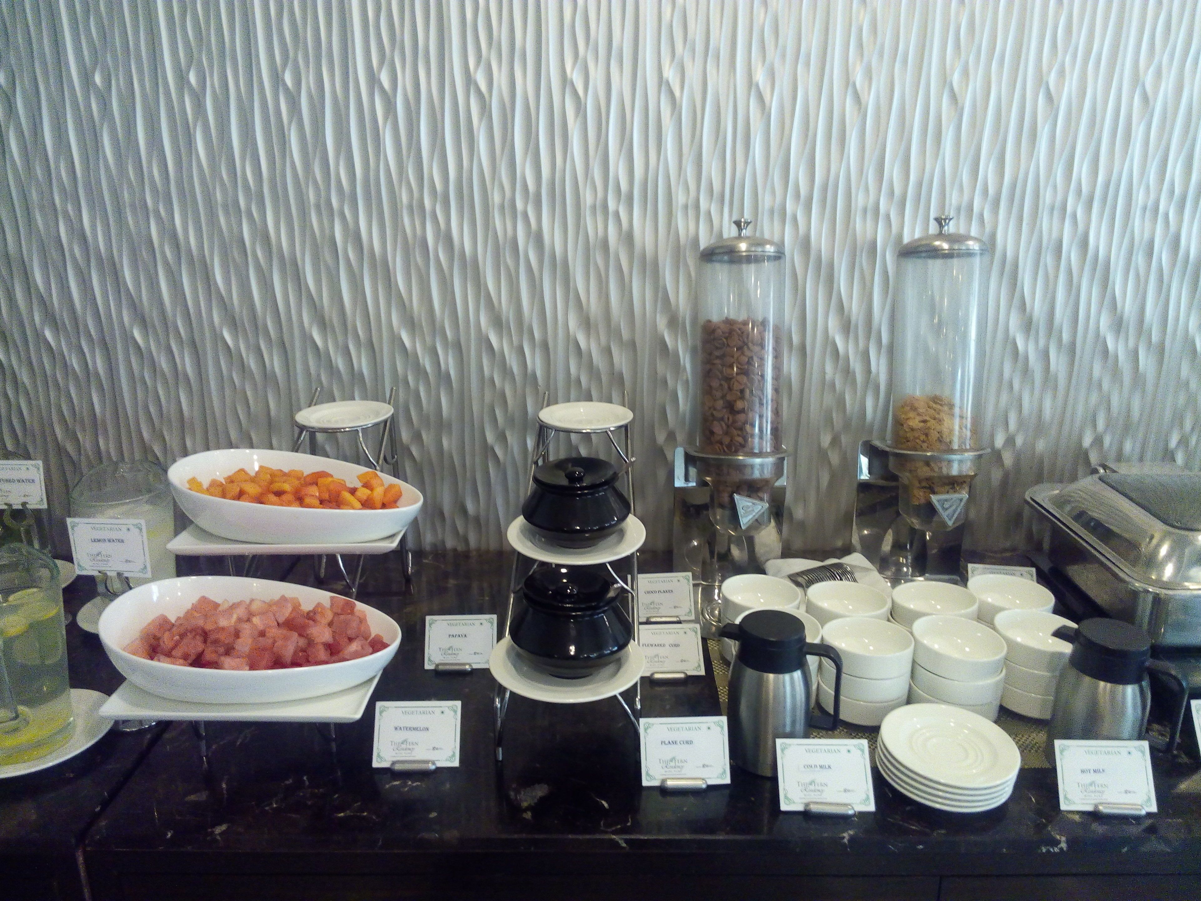 Buffet amb diversos plats i begudes, incloent bols de fruita, una estació de cafè i recipients etiquetats per diferents tipus de begudes.