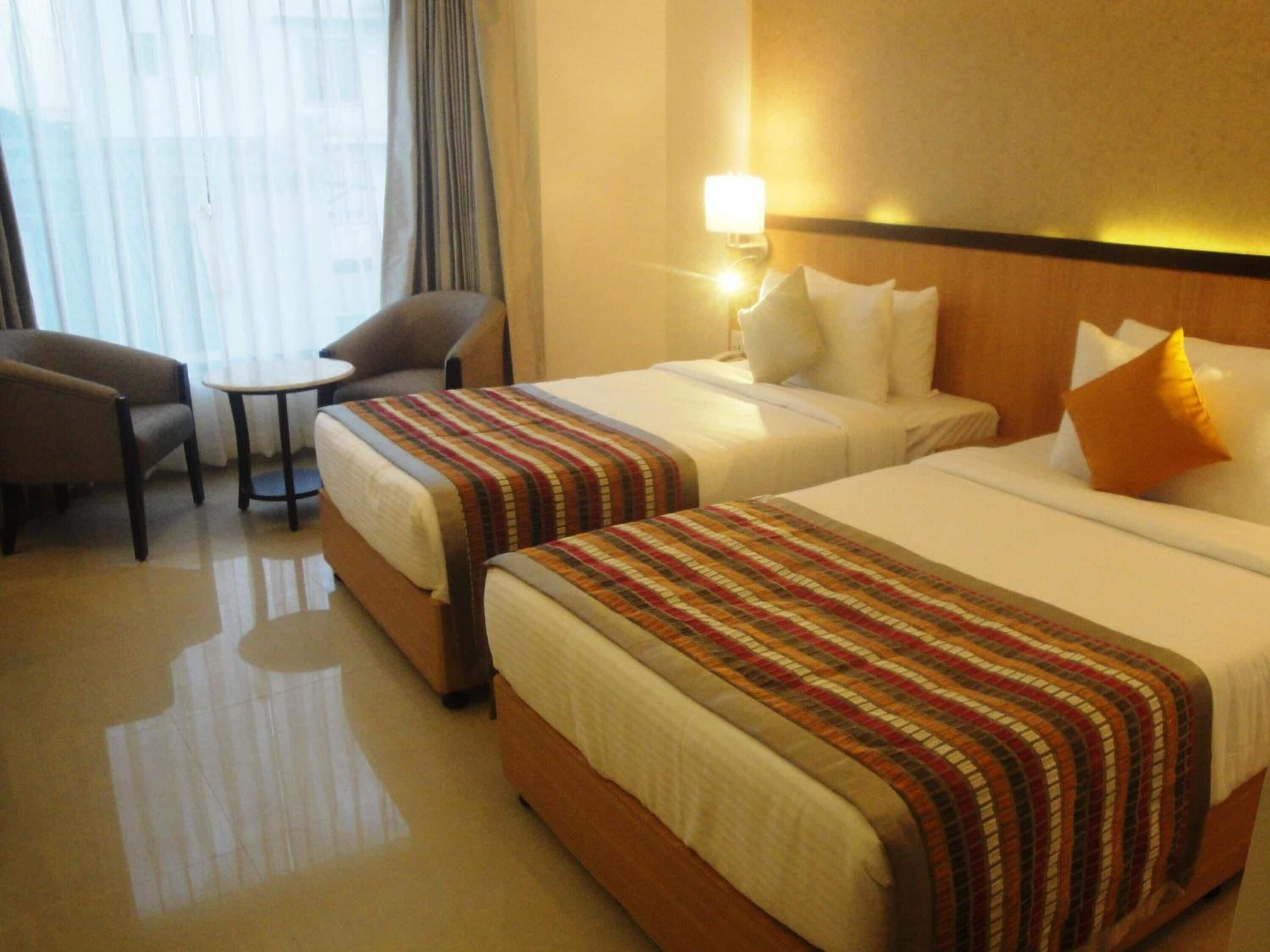 A hotel room with two beds, a small table, a chair, and a lamp.