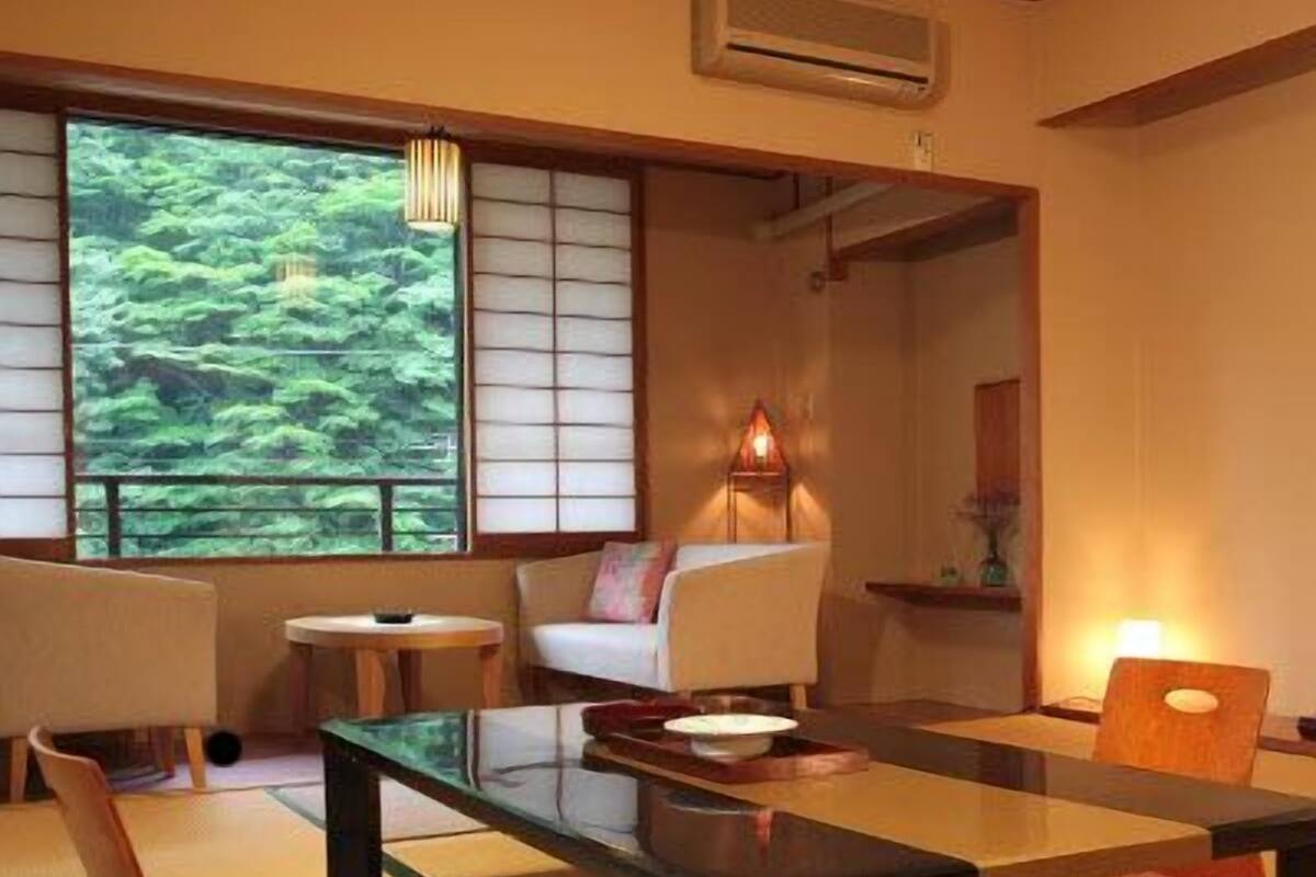 A traditional Japanese-style room with tatami flooring, a low table, a sofa, and a view of a forest through a shoji screen.