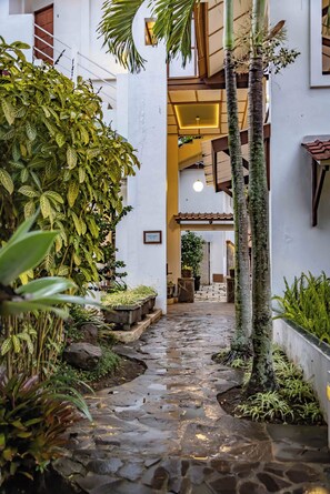 A narrow stone pathway flanked by lush greenery and white walls, leading to a building entrance with a wooden door and a sign. - Hotel Gradia 1 (Batu)