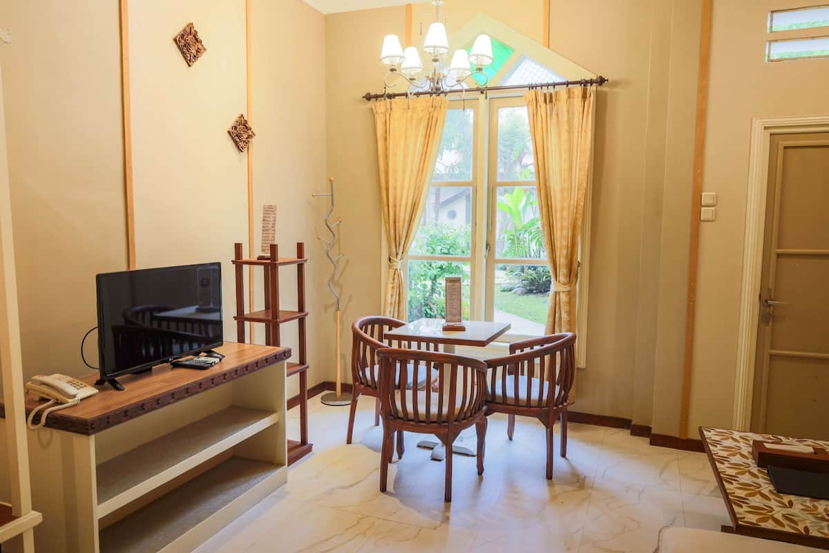 A room with a TV on a wooden stand, a dining table with chairs, a chandelier, and a window with curtains.