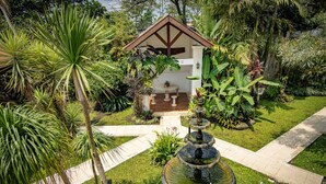 Ein Garten mit einem Brunnen, ein kleines weißes Gebäude mit braunem Dach und verschiedenen tropischen Pflanzen.