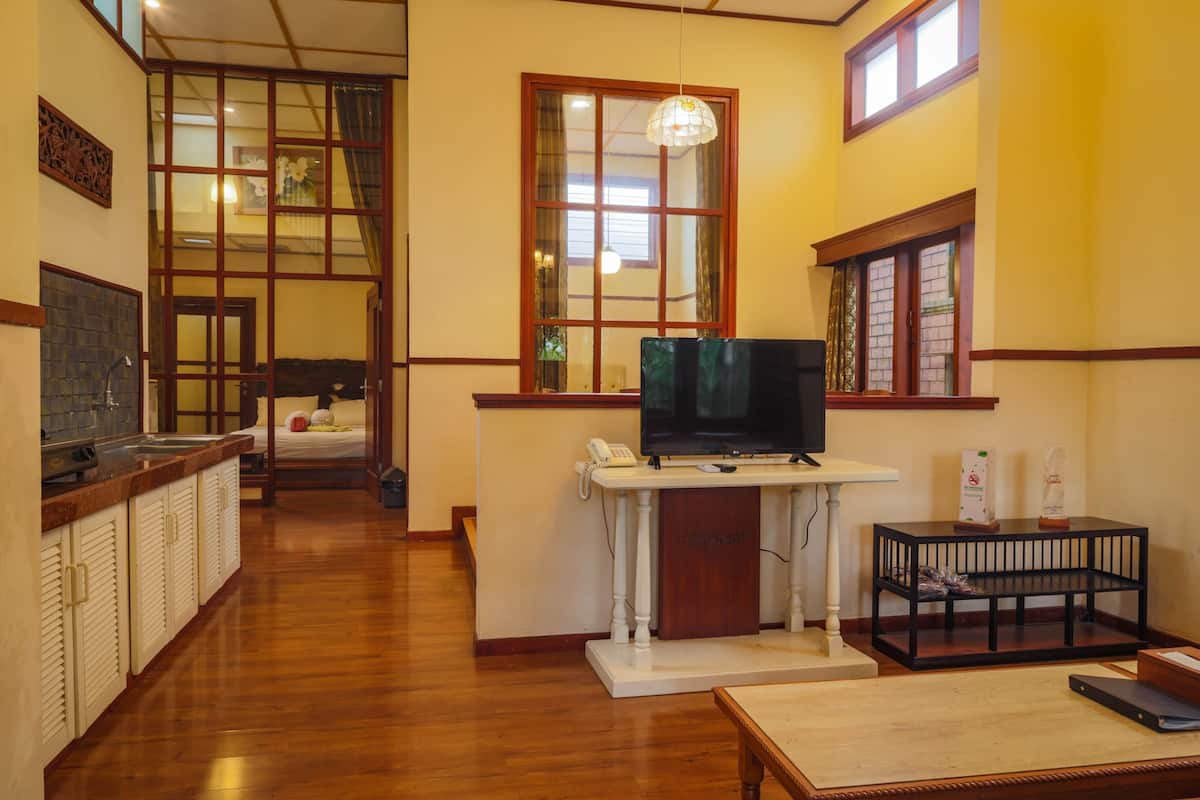 A hotel room with a wooden floor, a TV on a white stand, a small table, a window with curtains, and a view of the outside.