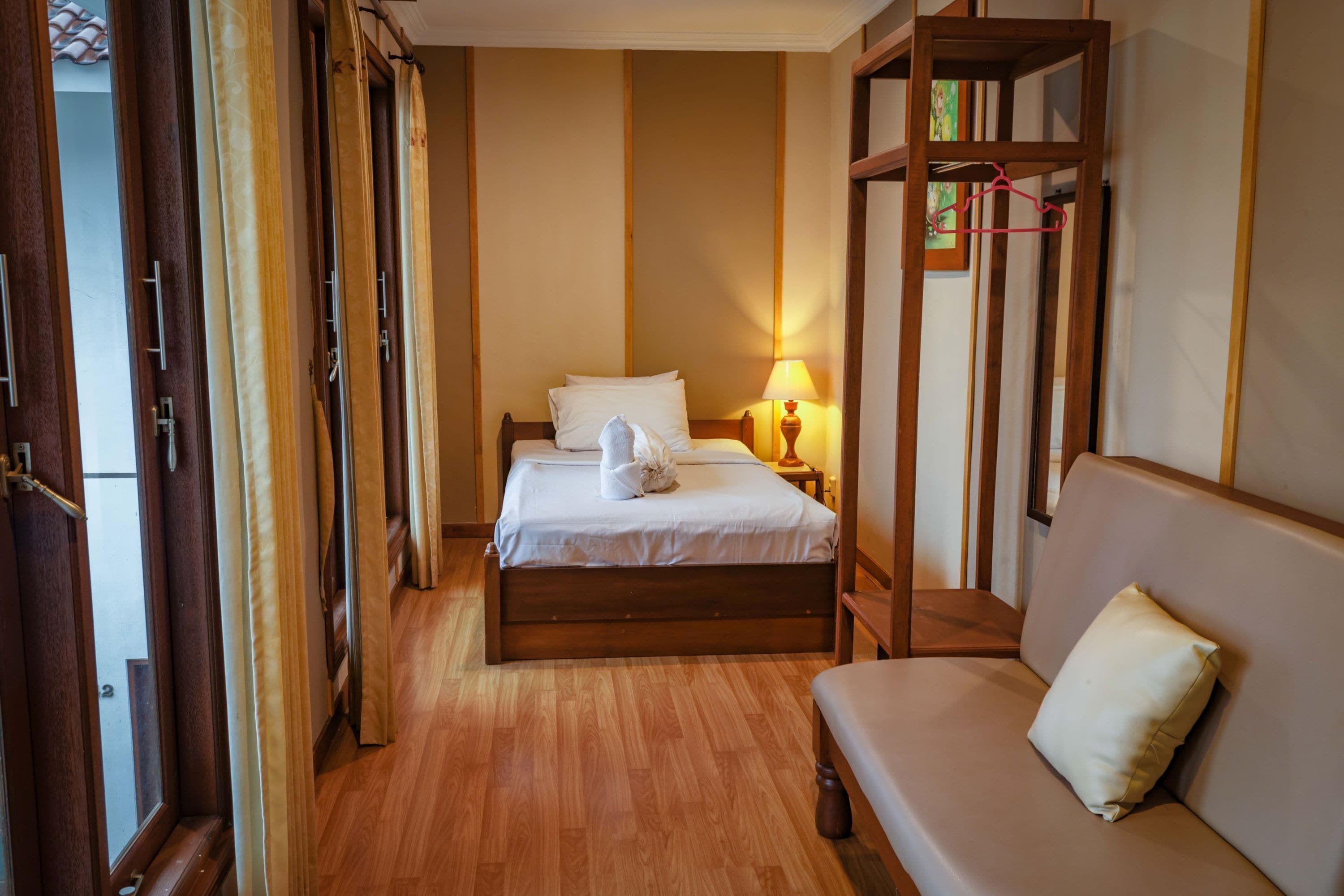 A hotel room with a wooden bed, a sofa, a nightstand with a lamp, and a wardrobe with a mirror.