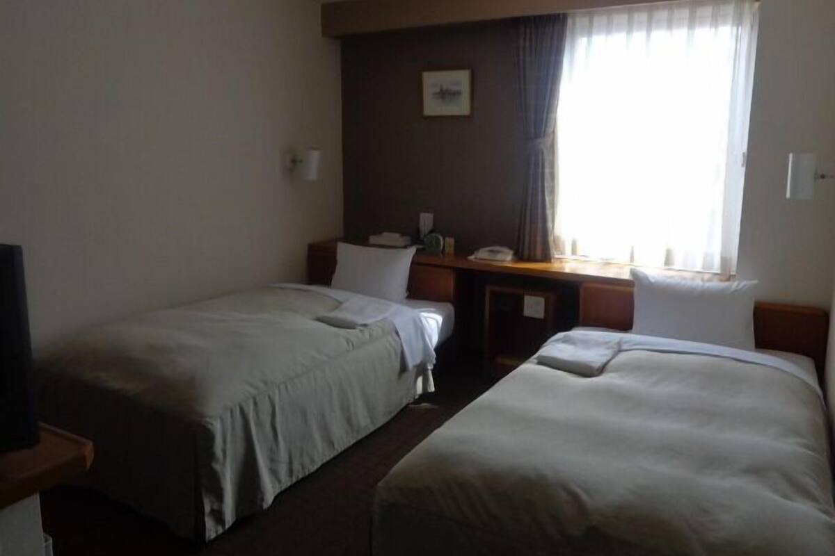 A hotel room with two single beds, a desk, a chair, and a window with curtains.
