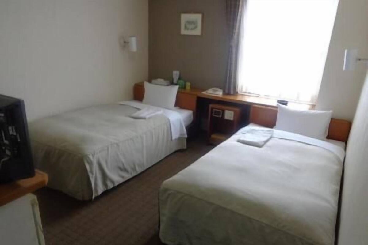 A hotel room with two beds, a desk, a chair, a small table, a television, and a window with curtains.