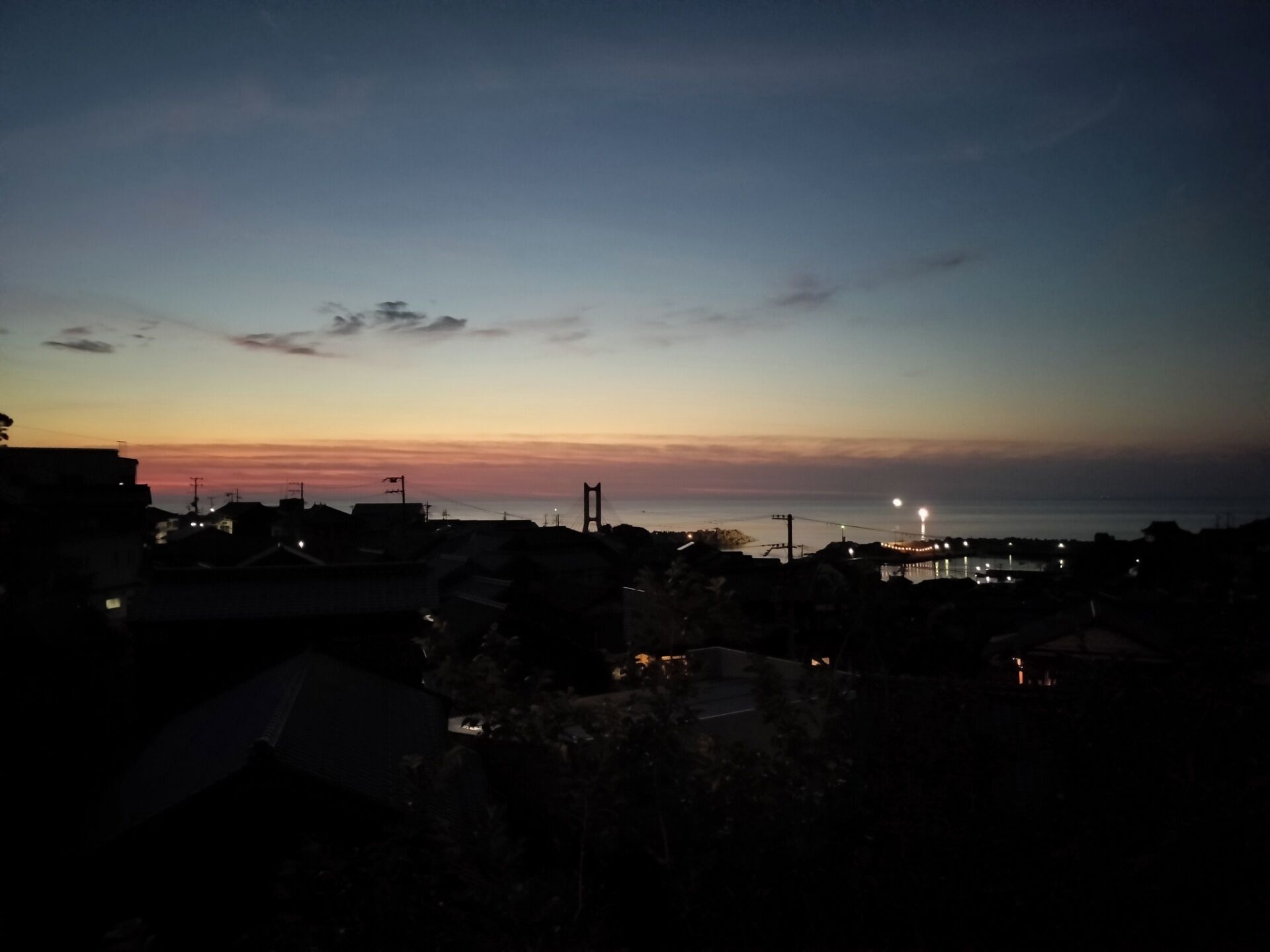 A coastal cityscape at dusk with silhouettes of buildings and a bridge.