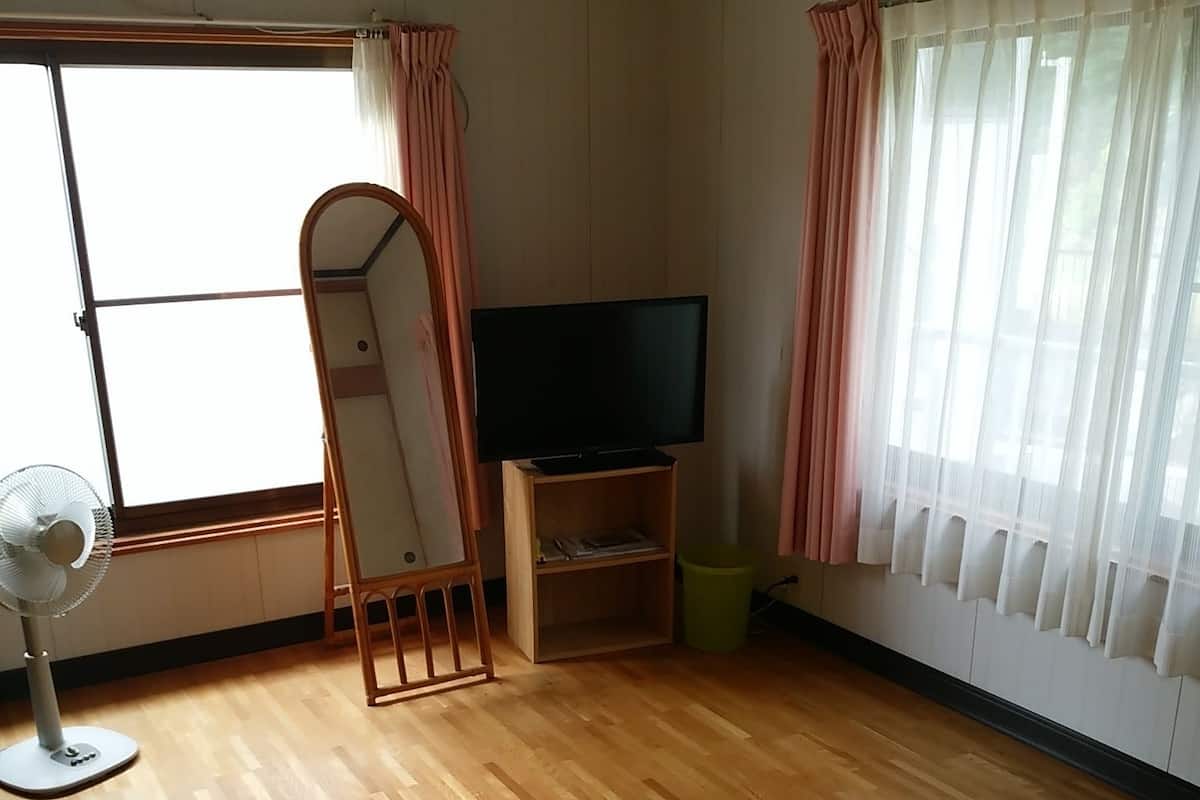 Ein Zimmer mit Holzboden, einem Stehspiegel, einem Flachbildfernseher auf einem Holzständer, einem Ventilator und durchsichtigen Vorhängen an den Fenstern.
