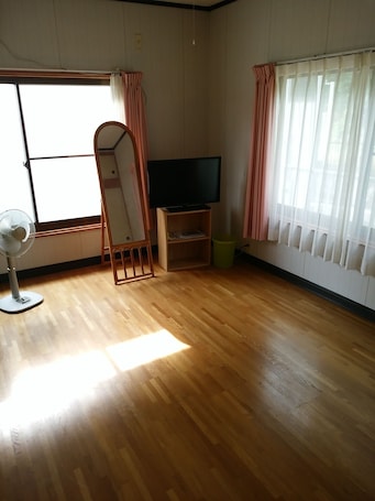A room with wooden flooring, a standing mirror, a flat-screen TV on a wooden stand, a fan, and sheer curtains on the windows.