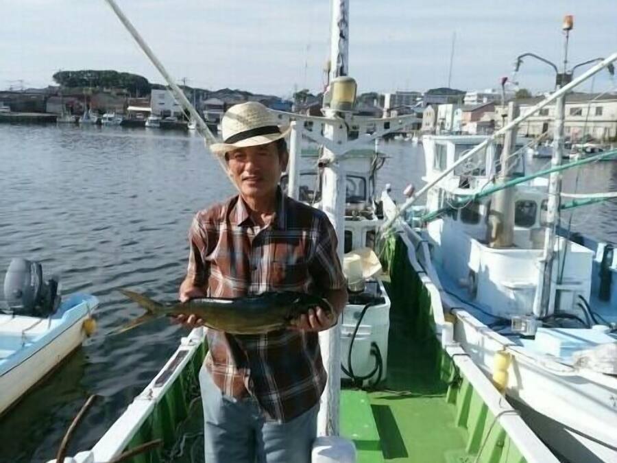 Iemand in een boot met een vis, met andere boten en een jachthaven op de achtergrond.