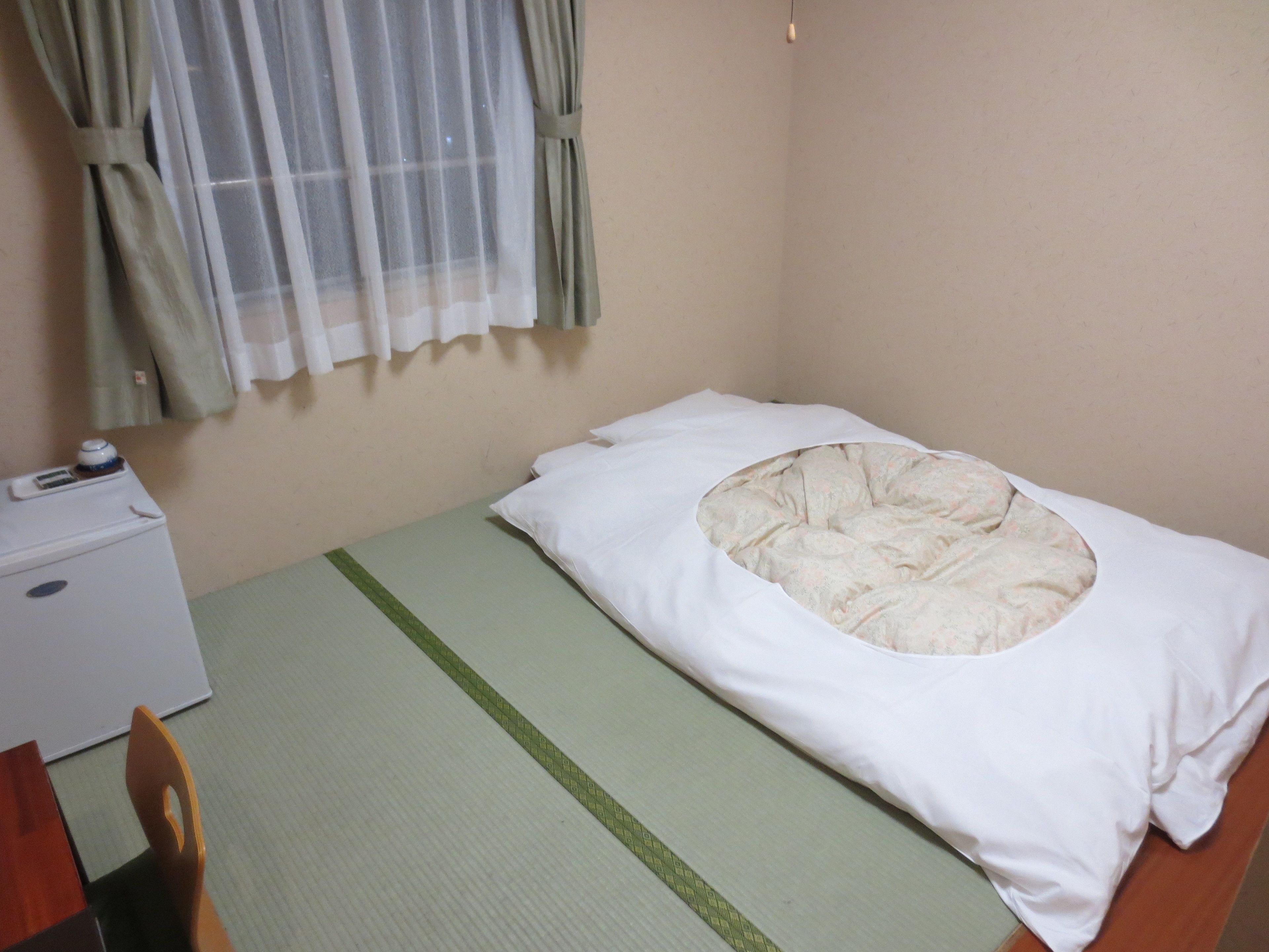 Una habitación pequeña y sencilla con una cama, una silla, un escritorio con un ordenador y una ventana con cortinas.