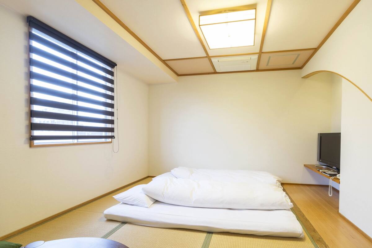 Una habitación pequeña y ordenada con una cama, un televisor y una ventana con persianas.