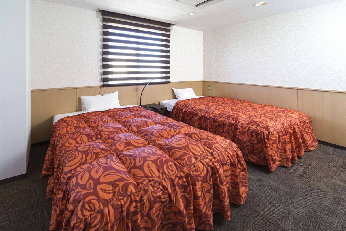 Una habitación de hotel con dos camas, un ventanal con persianas y suelo alfombrado.