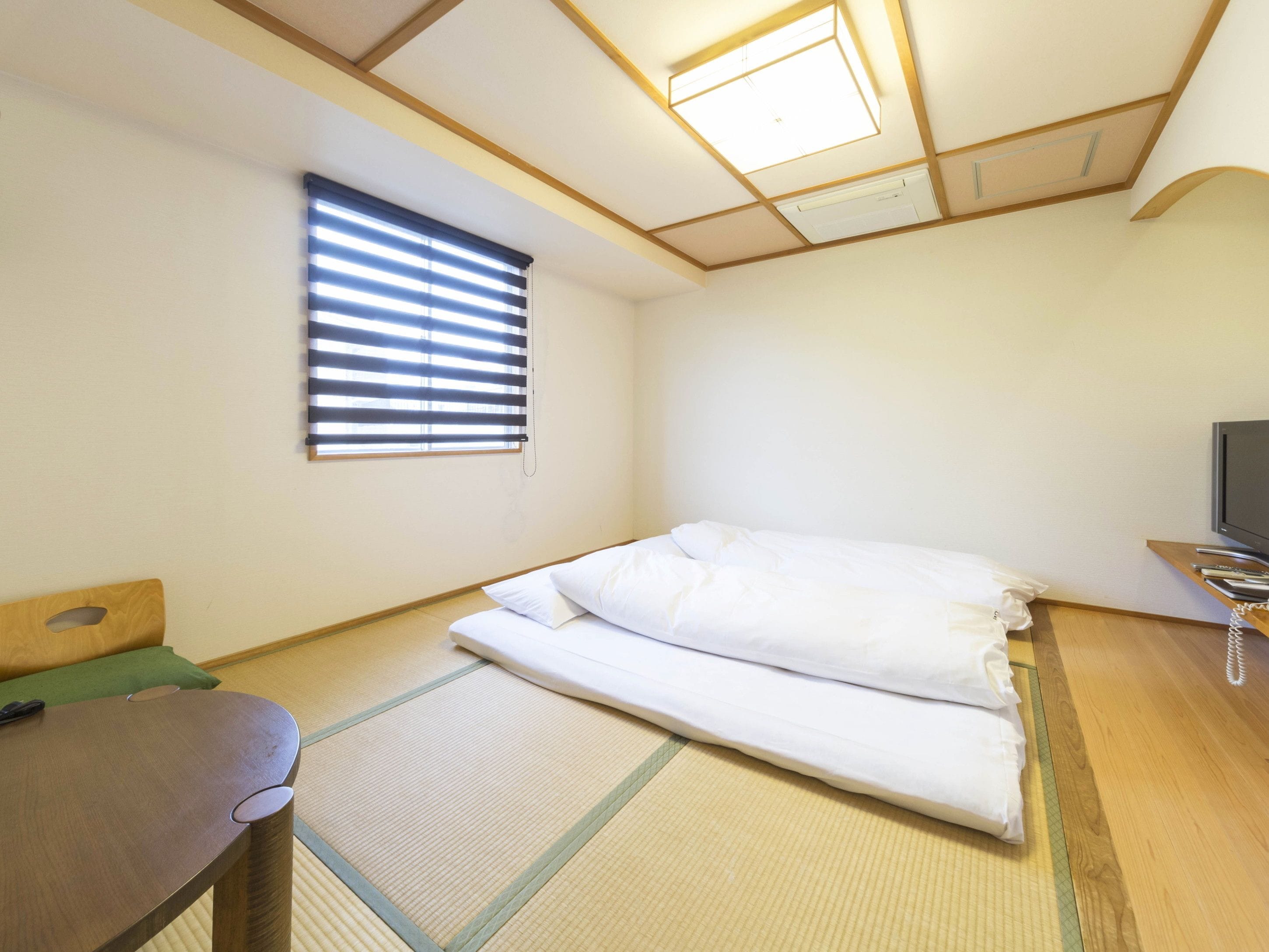 A minimalist room with tatami flooring, a bed with white bedding, a small wooden table, and a television on a shelf.
