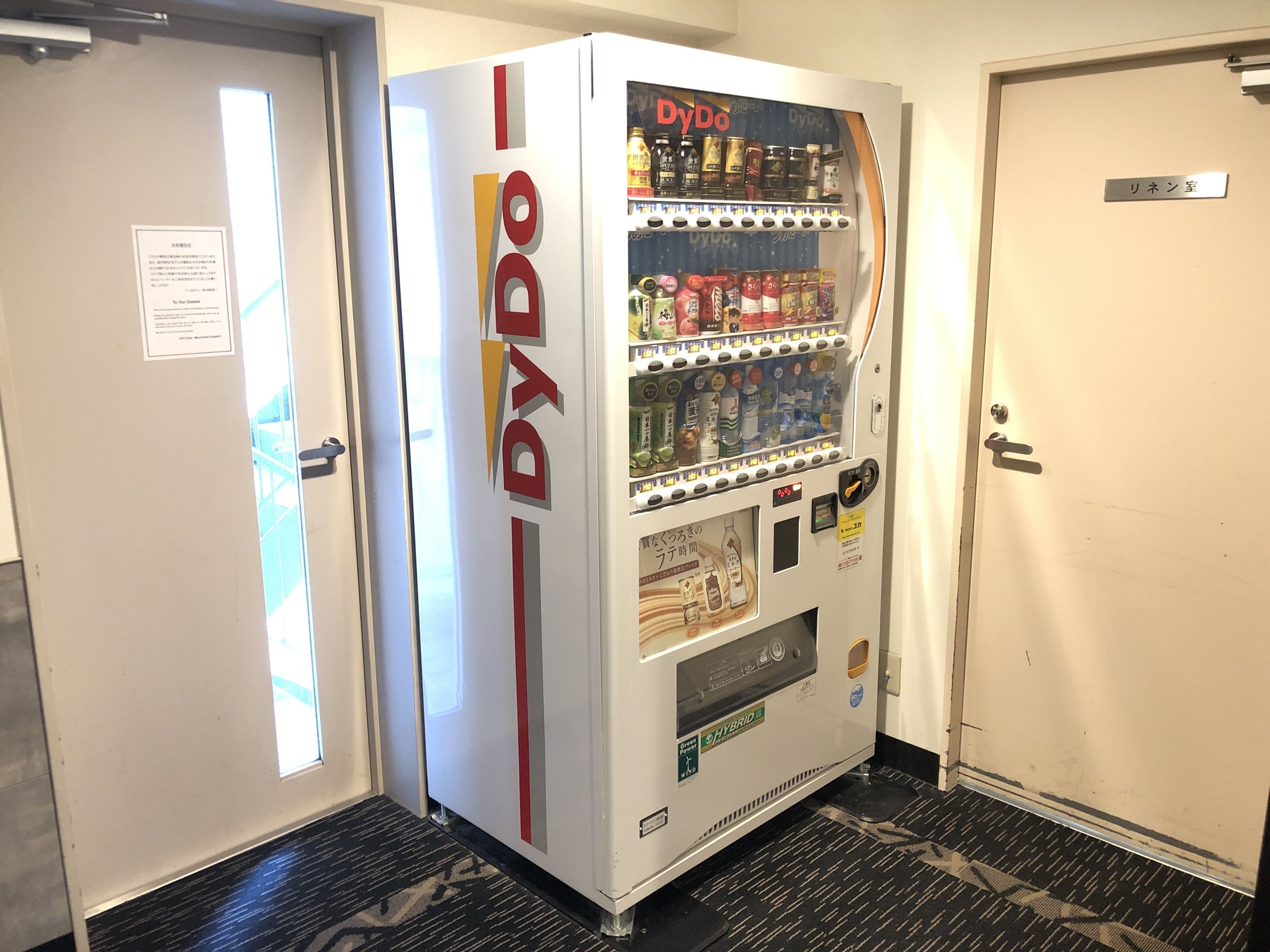 A vending machine labeled 'DyDo' in a hotel corridor with a door labeled 'リネン室'.