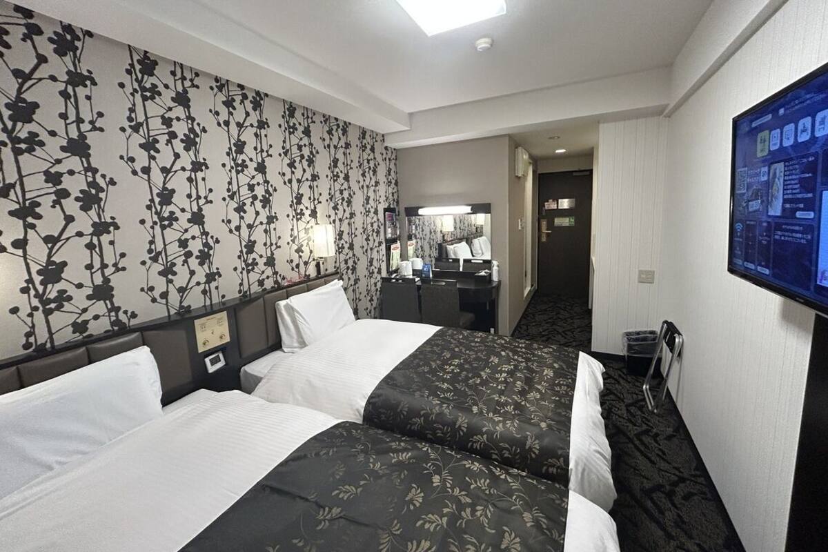 A hotel room with two beds, a television, a desk with a chair, and a bathroom visible through an open door.