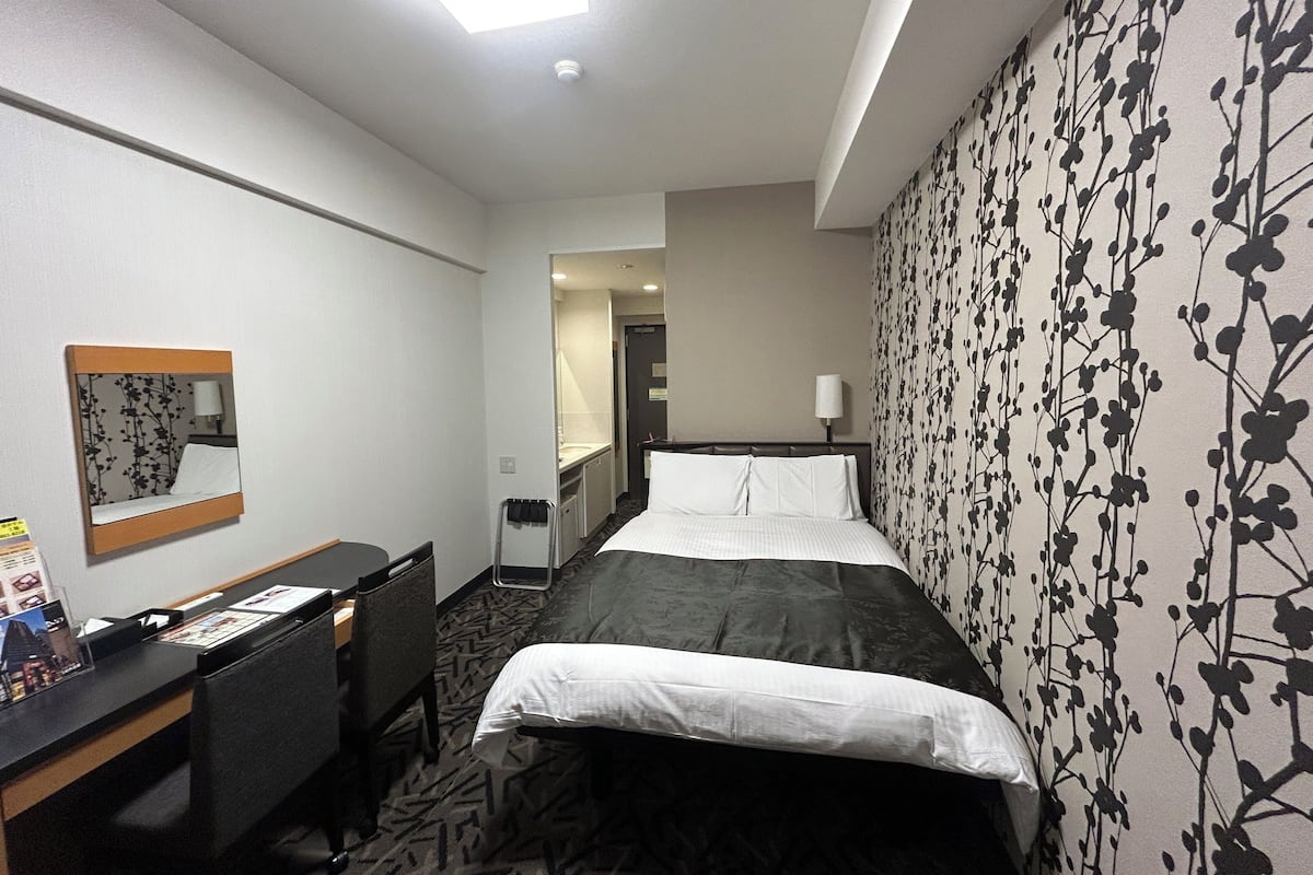 A hotel room with a bed, a desk, a chair, and a mirror.