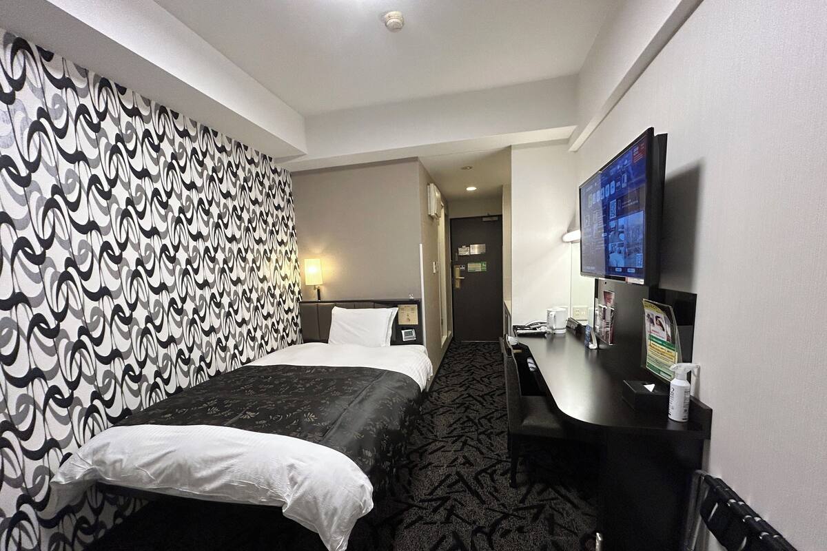A hotel room with a patterned wall, a bed with white linens, a desk with a television, and a carpeted floor.