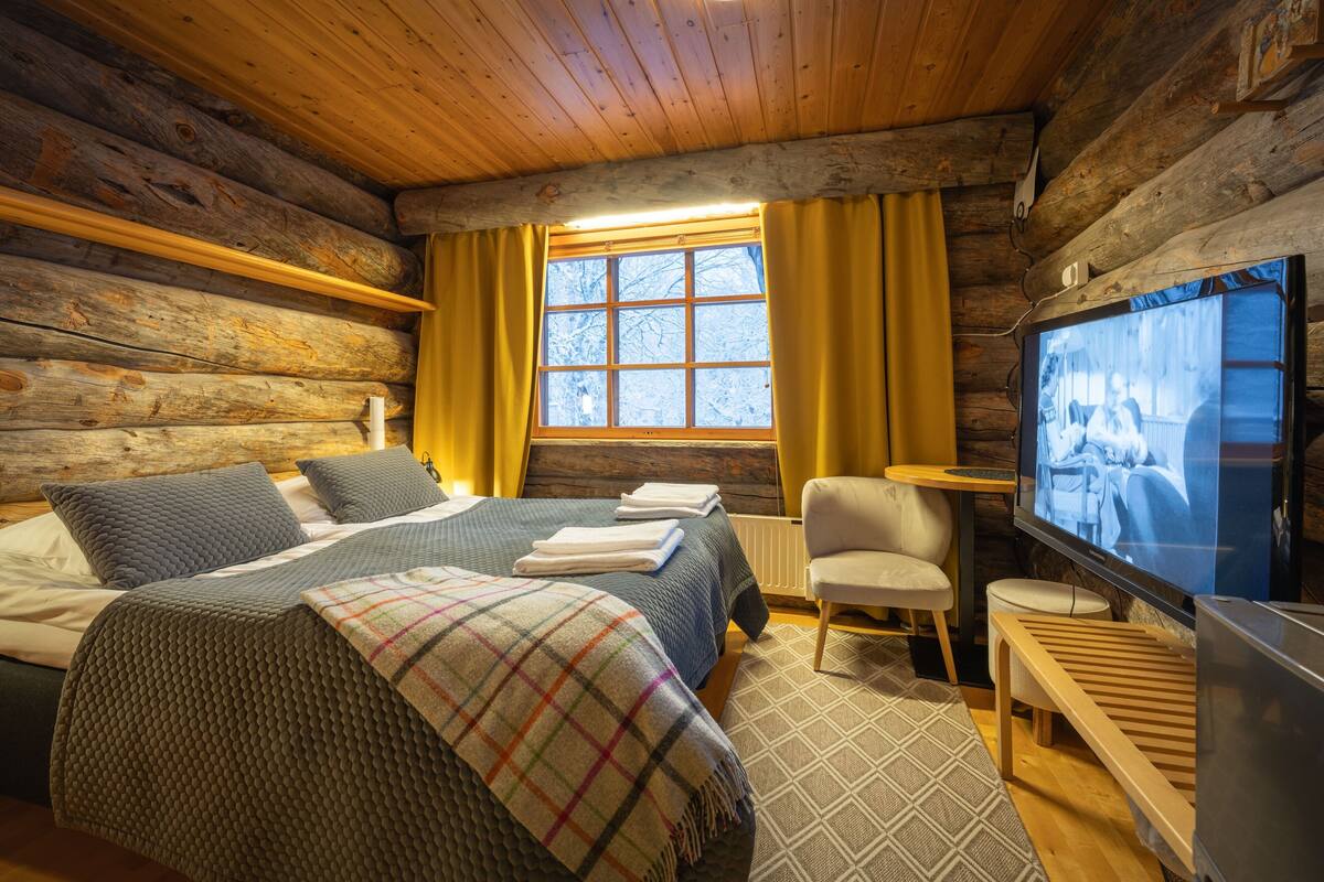 Une chambre aménagée dans un chalet en rondins, avec un lit, un téléviseur, un banc en bois et une fenêtre avec des rideaux.
