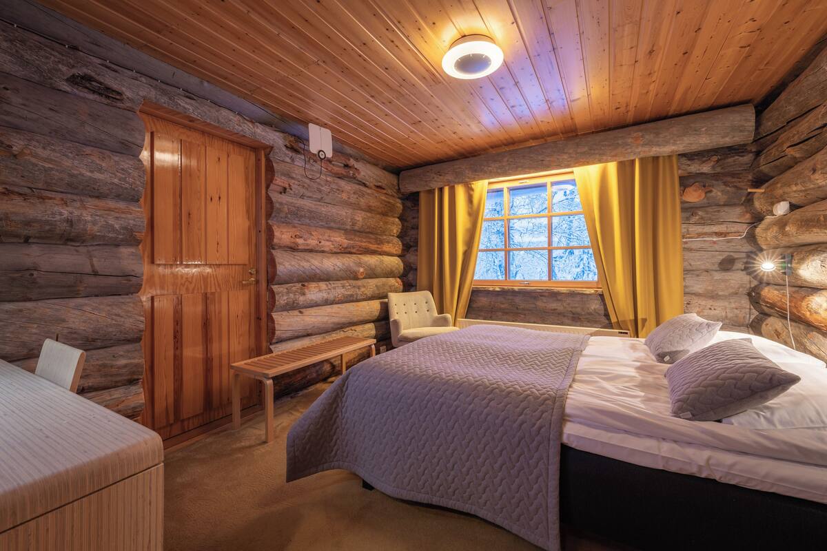 Une chambre dans un chalet en rondins, avec un lit, une chaise, une table et une fenêtre avec des rideaux.