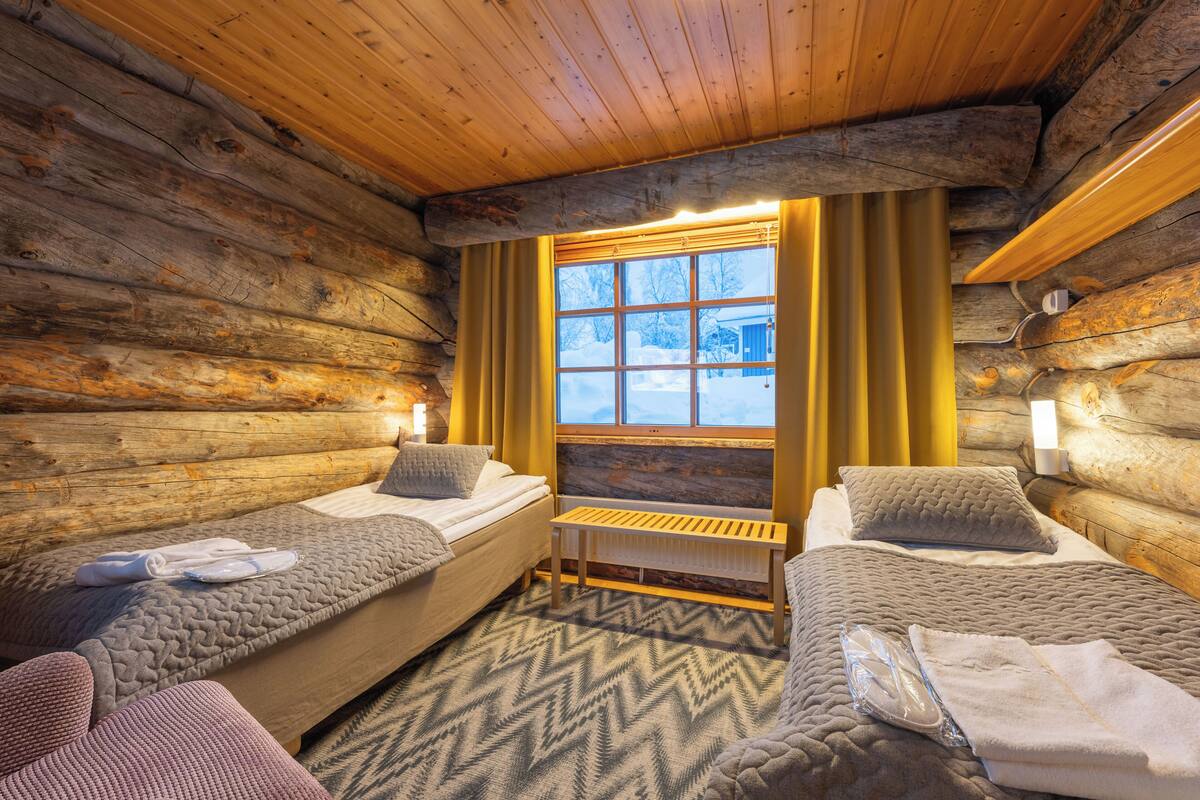 Une chambre aménagée dans un chalet en rondins, avec deux lits, un banc et une fenêtre avec des rideaux.