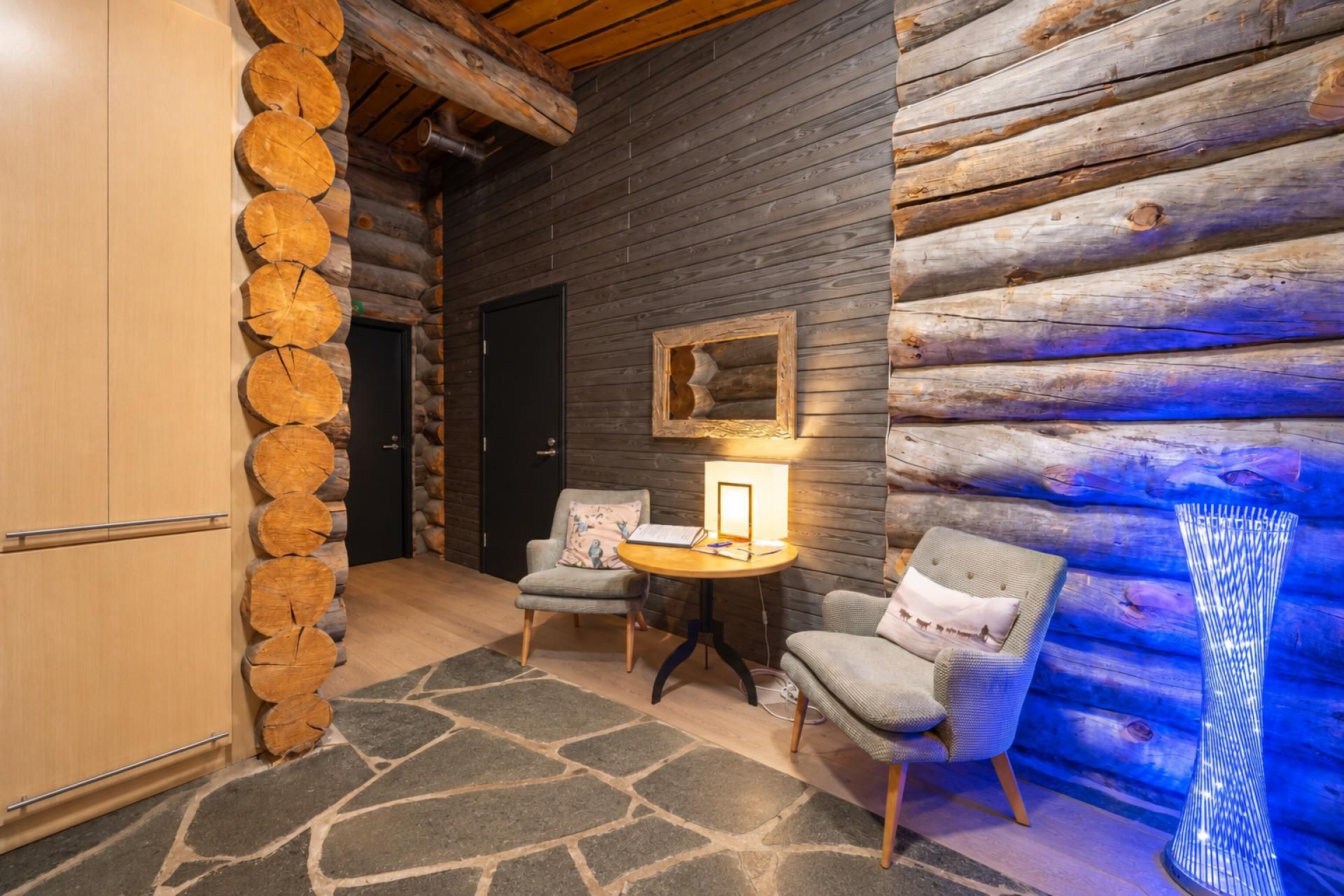 A log cabin interior with a stone wall, a round table with a lamp, two chairs, and a wooden cabinet.