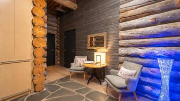 A log cabin interior with a stone wall, a round table with a lamp, two chairs, and a wooden cabinet.