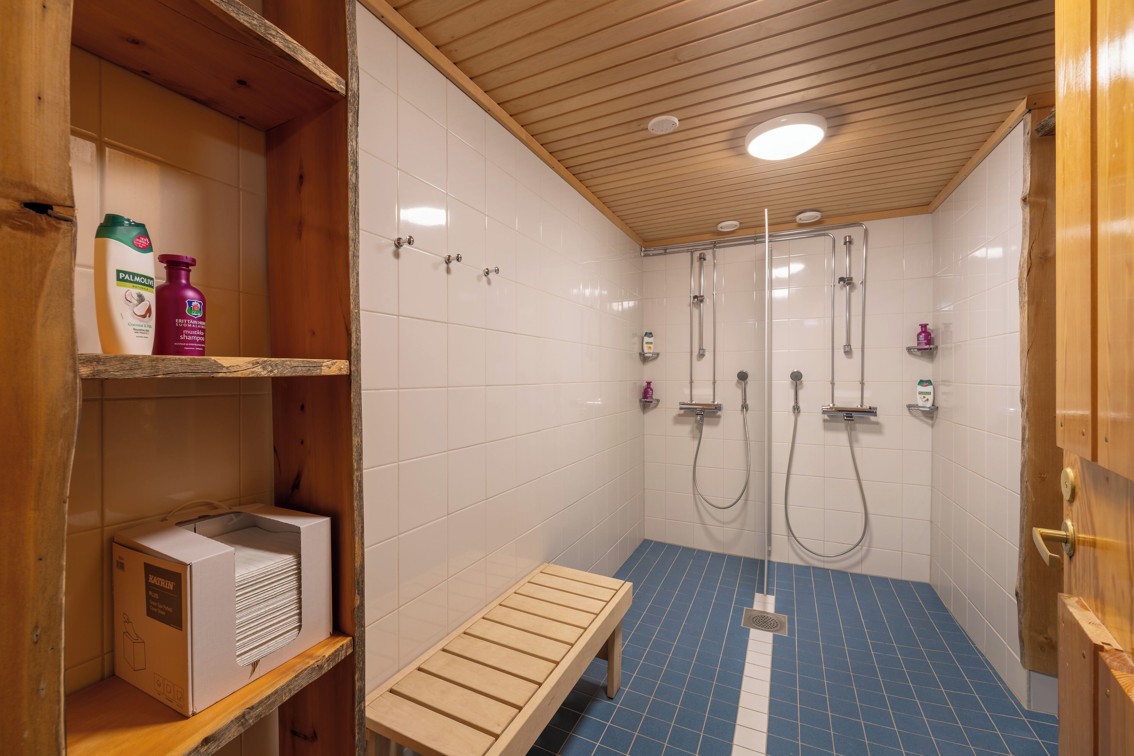 A shower area with two glass-enclosed showerheads, a bench, and shelves with toiletries.