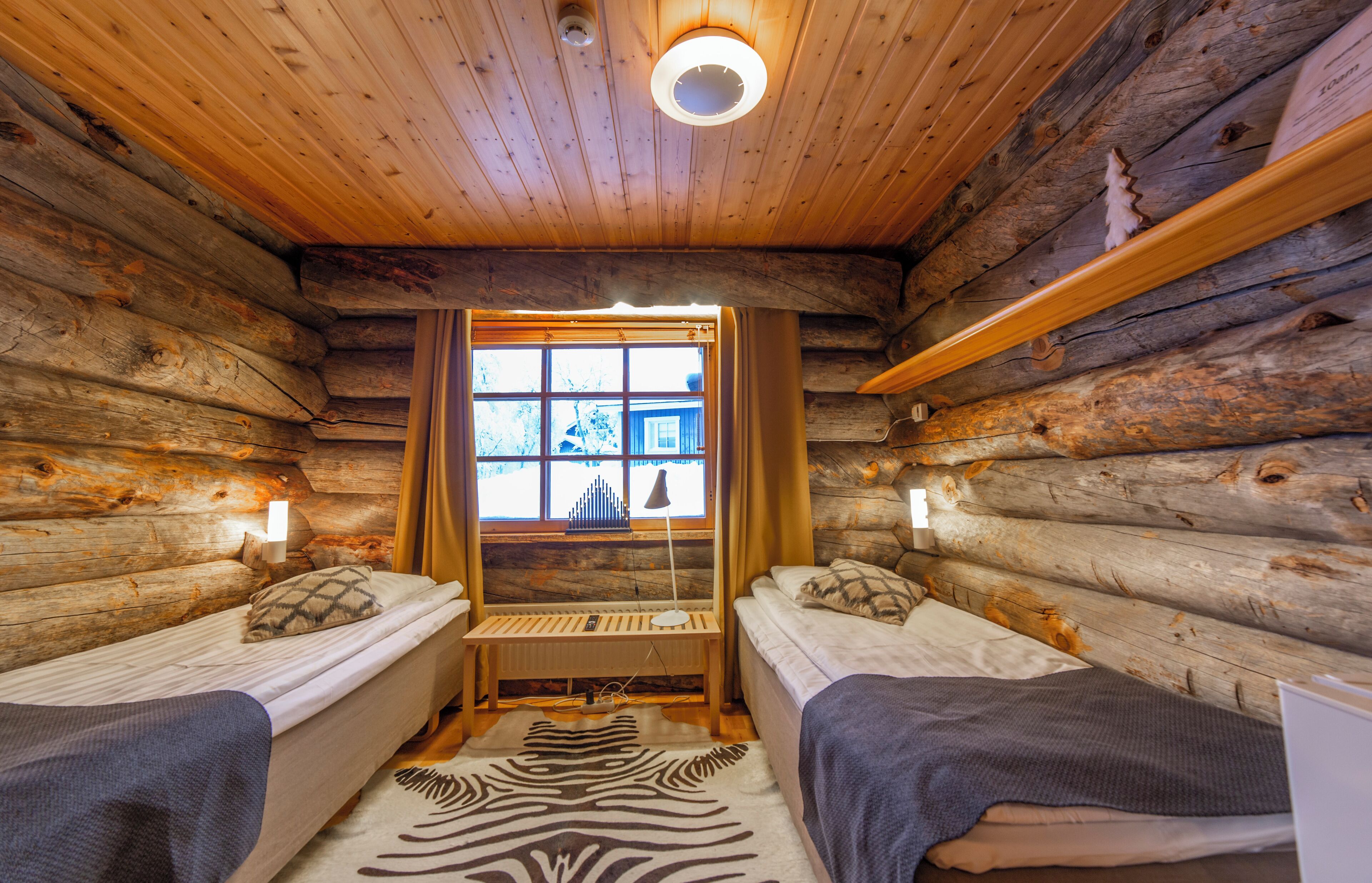 A log cabin room with two beds, a zebra-patterned rug, a window with curtains, and a wooden ceiling.