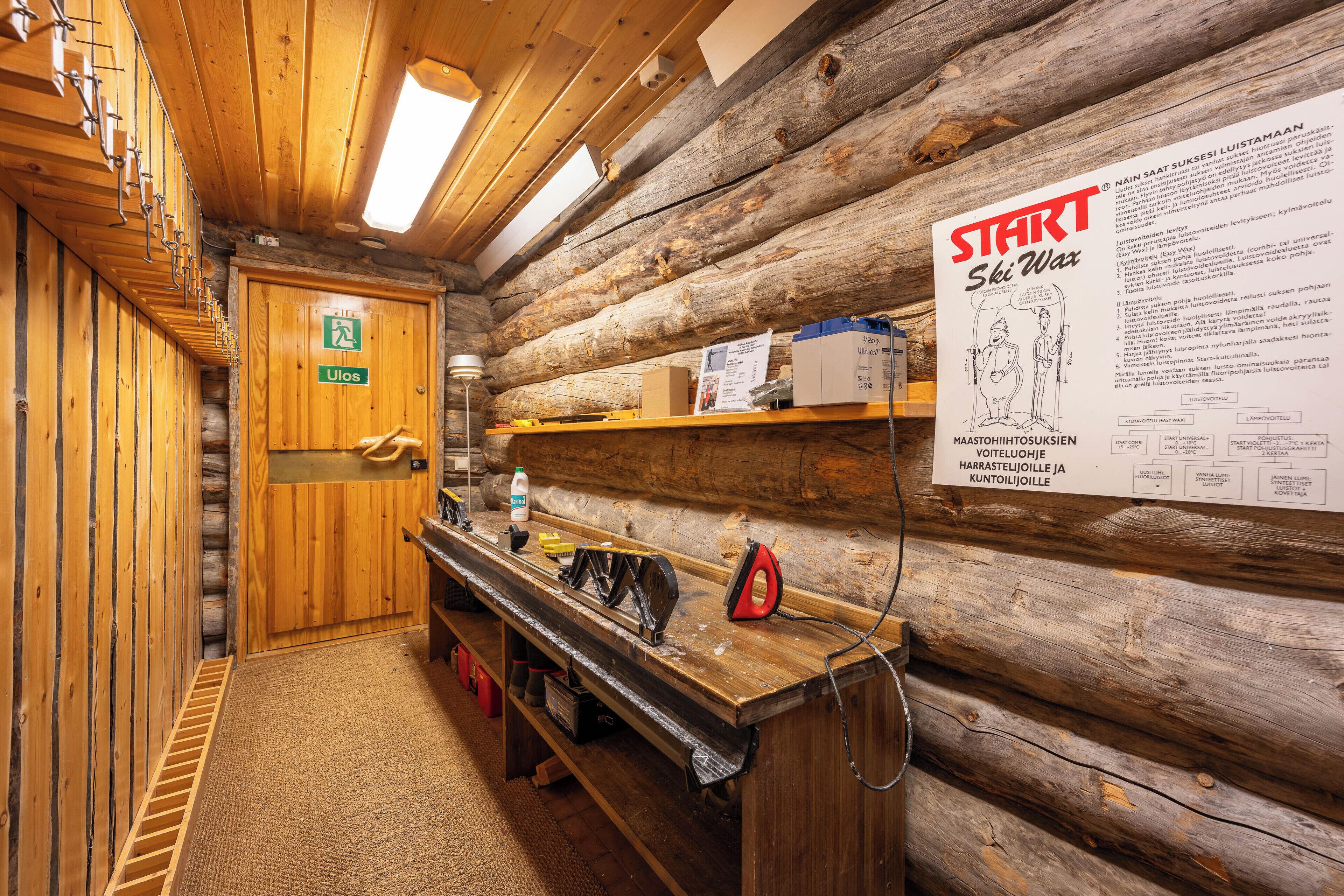A wooden cabin interior with a wooden door marked "Ulos," a wooden bench, a shelf with items, and a poster titled "START Ski Wax."