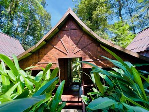 A traditional wooden structure with a thatched roof, surrounded by lush greenery.