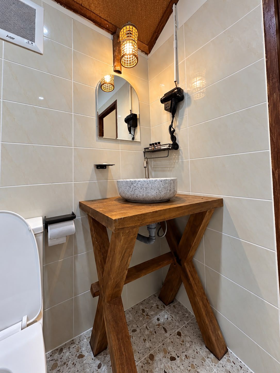A bathroom with a white tiled wall, a wooden vanity with a bowl sink, a hairdryer mounted on the wall, and a toilet with a paper towel holder.