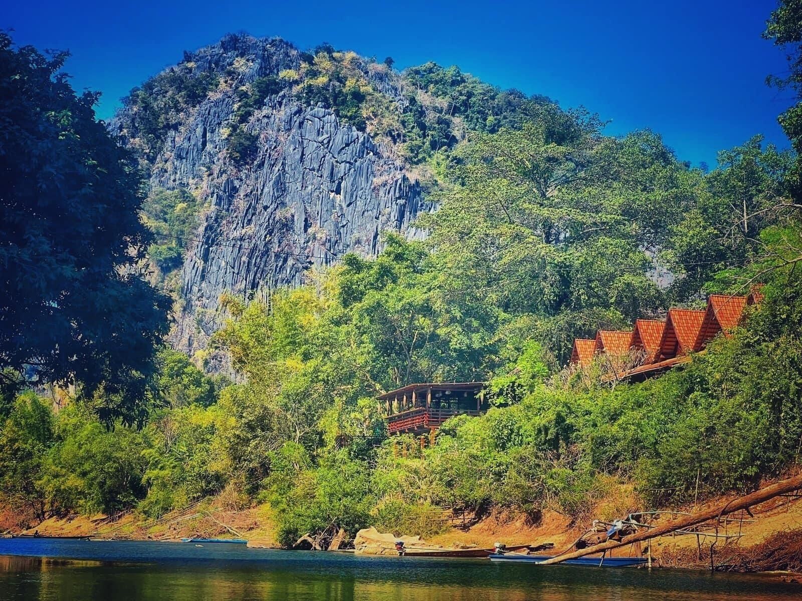 A traditional wooden structure with a thatched roof, surrounded by lush greenery and a rocky cliff.