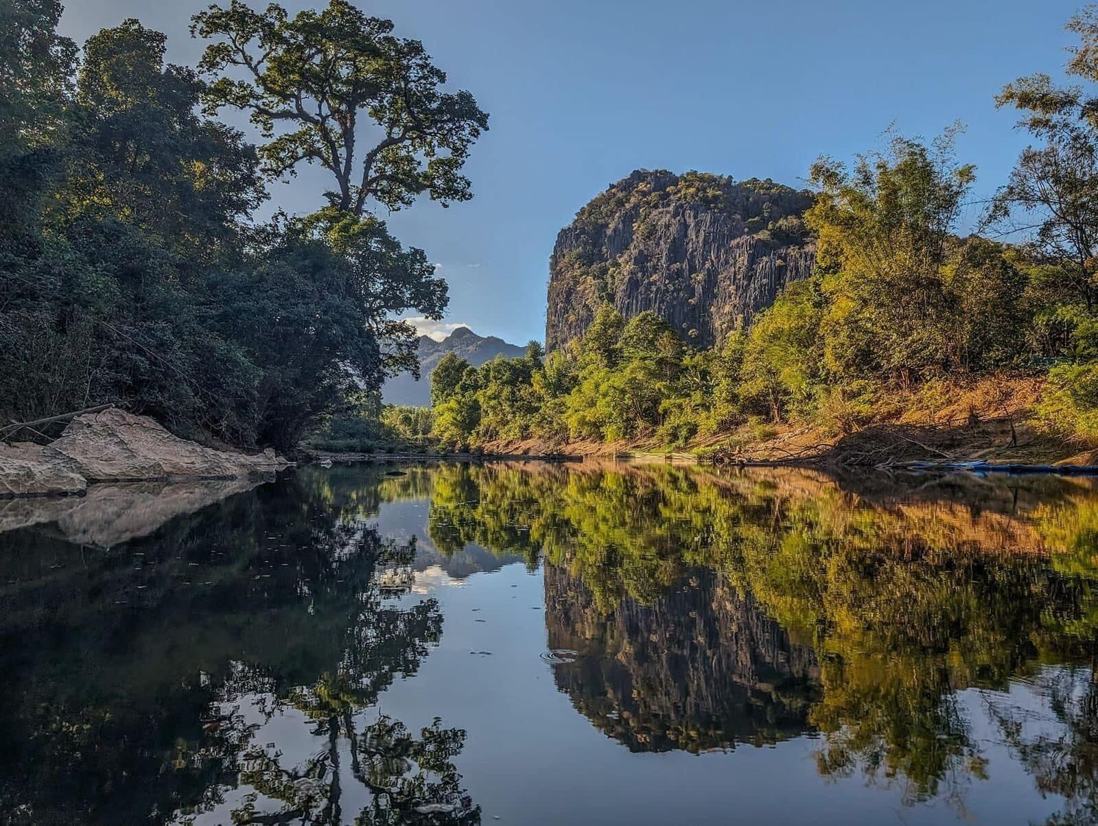 A calm river reflecting trees and a rocky mountain.