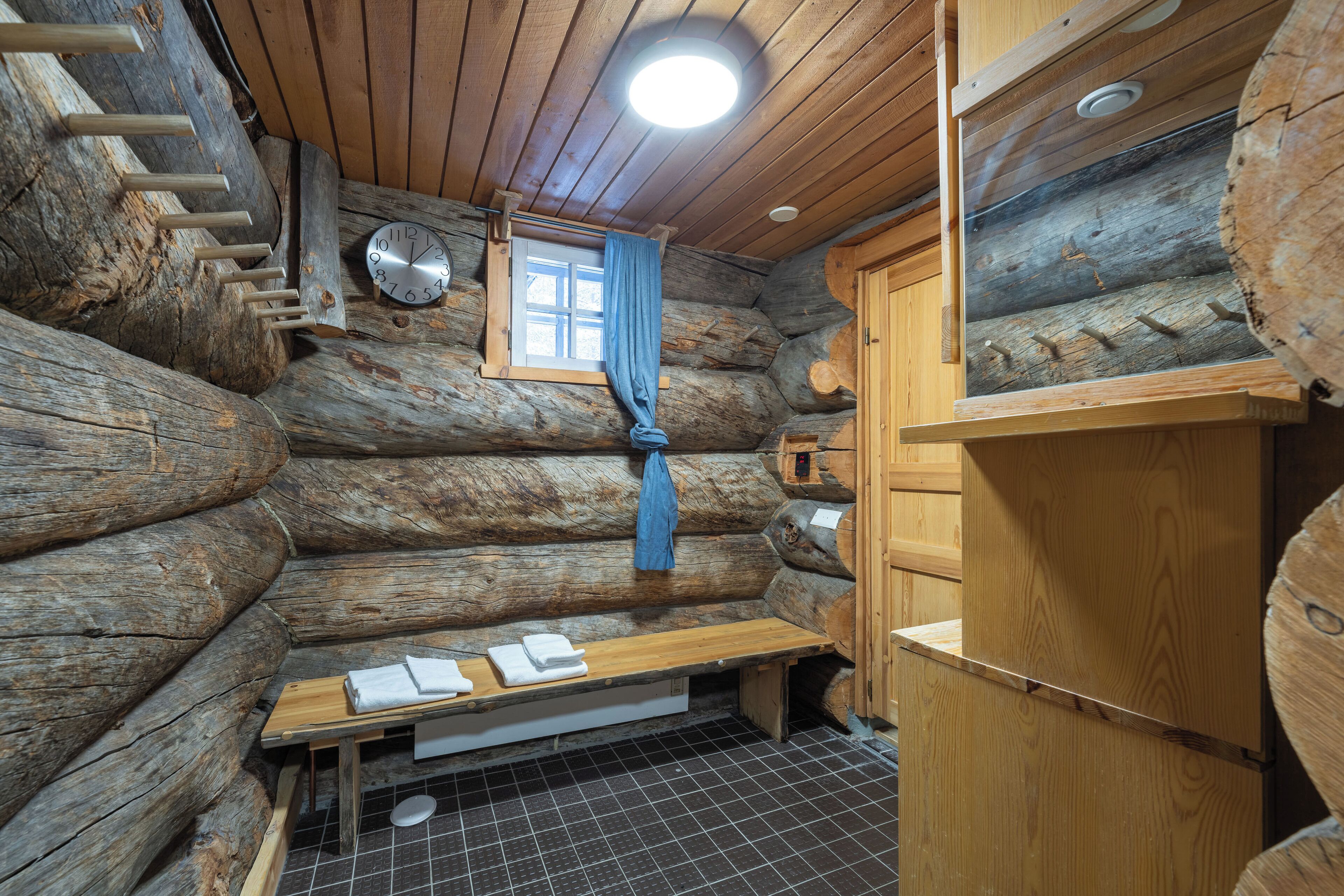 A log cabin interior with a wooden bench, a small window with a curtain, and a wooden cabinet.