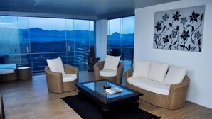 Una sala de estar moderna con muebles de mimbre, una mesa de centro de vidrio y vistas a montañas a través de ventanas amplias.