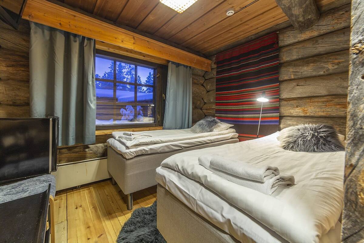 A cozy log cabin bedroom with two beds, a wooden ceiling, and a window overlooking a snowy landscape.
