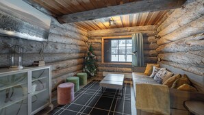 Interior de una cabaña de troncos con techo de madera, un aparador de cristal, un pequeño árbol de Navidad y una cama con almohadas.