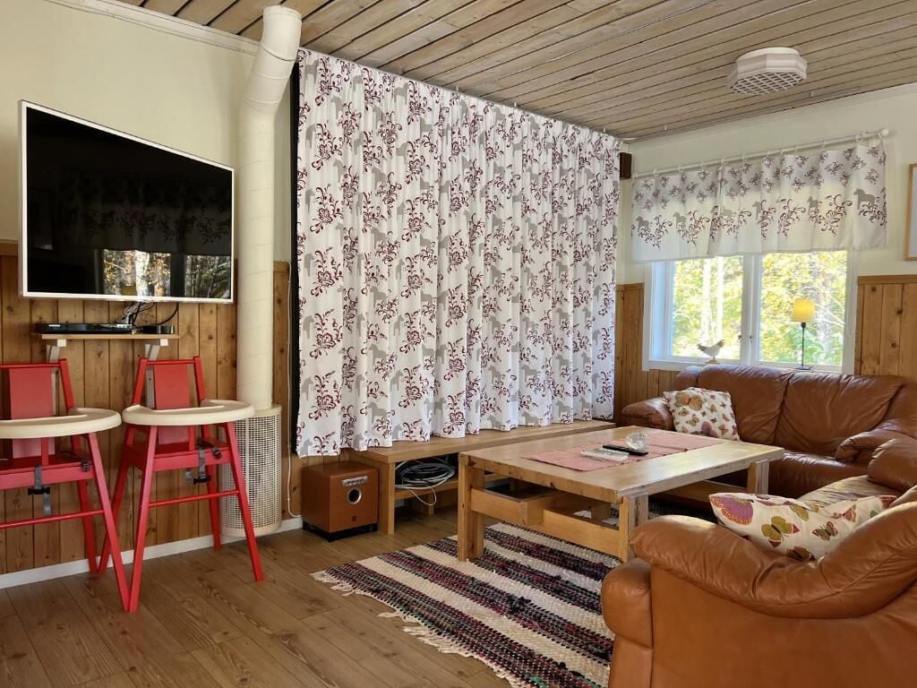 Una sala de estar con televisor, sillas rojas, un sofá marrón, una mesa de centro de madera y cortinas estampadas.