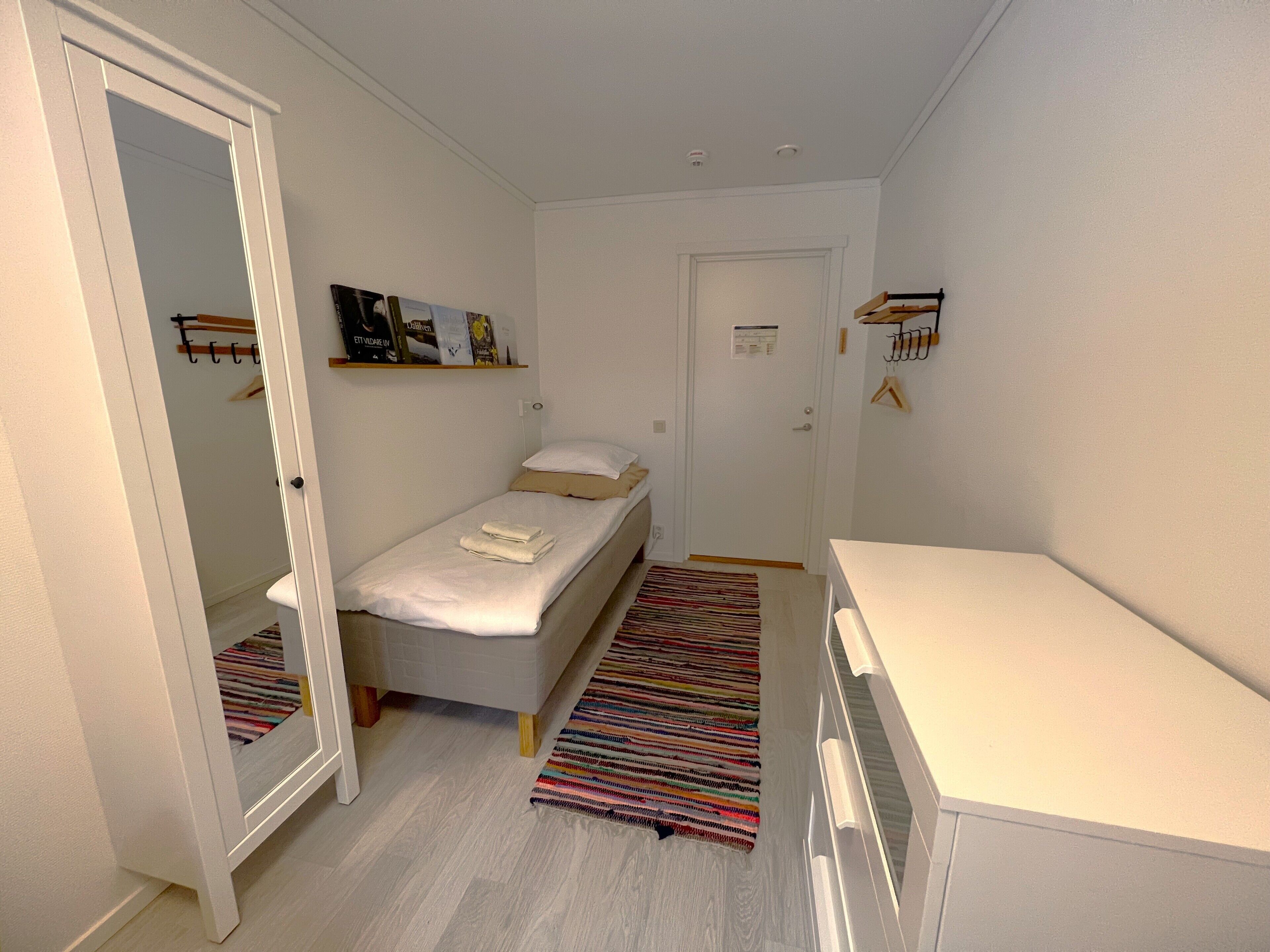 Ein kleines, helles Schlafzimmer mit einem Einzelbett, einem Nachttisch, einem Spiegel und einer Tür zu einem anderen Raum.