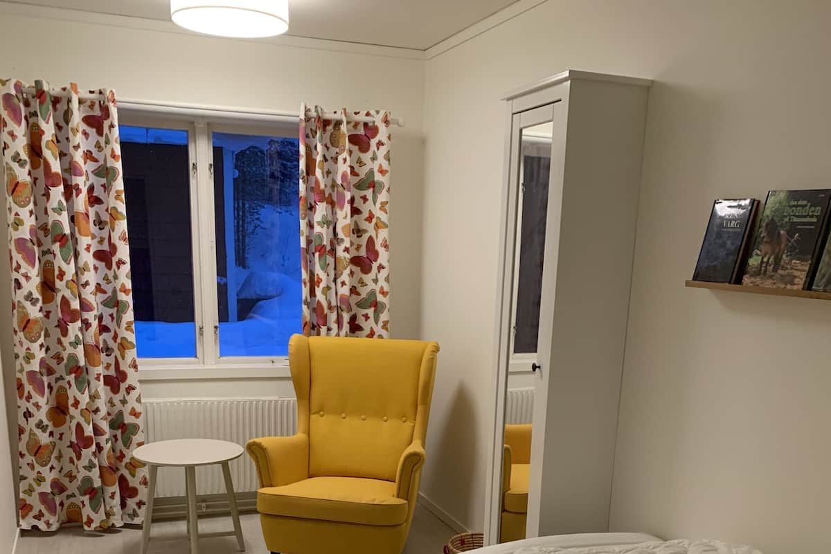 Een kamer met een bed, een gele fauteuil, een klein tafeltje, een kleurrijk vloerkleed en een raam met gordijnen.