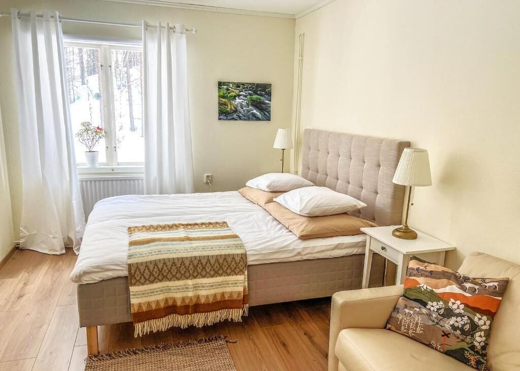 Un dormitorio con cama, lámparas de noche, una silla, ventana con cortinas, radiador y un cuadro en la pared.