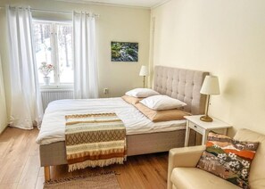 Un dormitorio con cama, lámparas de noche, una silla, ventana con cortinas, radiador y un cuadro en la pared.