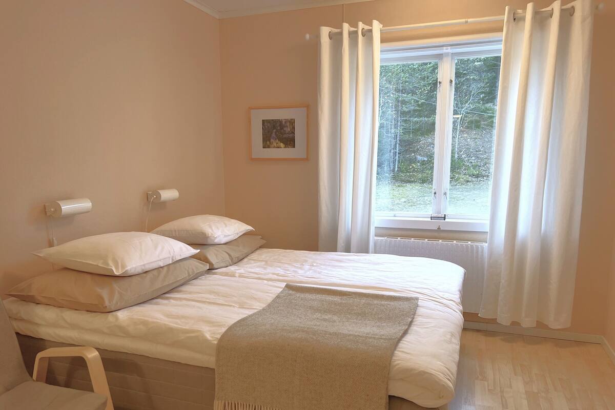 Een slaapkamer met een bed, een stoel, een raam met gordijnen, een radiator en een lijst met een afbeelding aan de muur.
