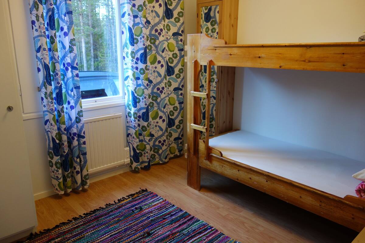 Een kamer met een stapelbed, een raam met gordijnen, een radiator en een kleurrijk tapijt.