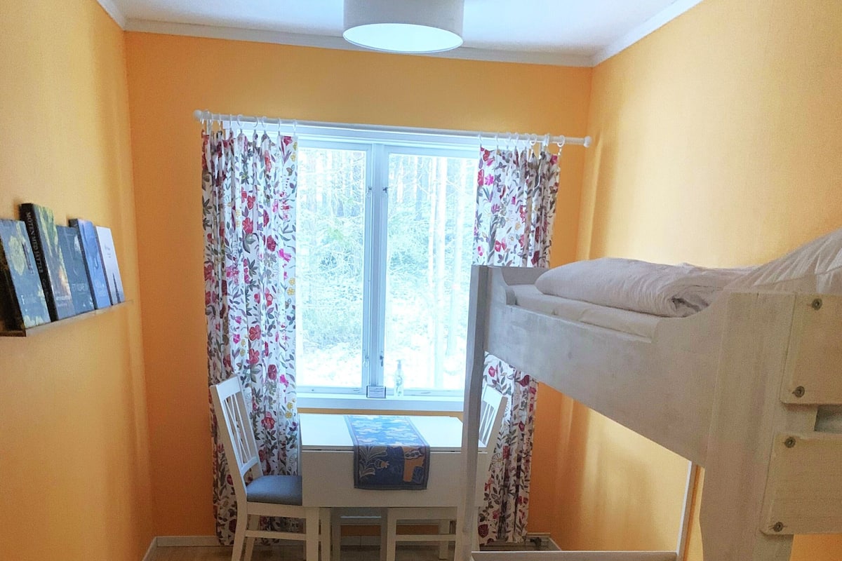 Een kleine kamer met een stapelbed, een bureau met een stoel, een raam met bloemen gordijnen en een kleurrijk vloerkleed.