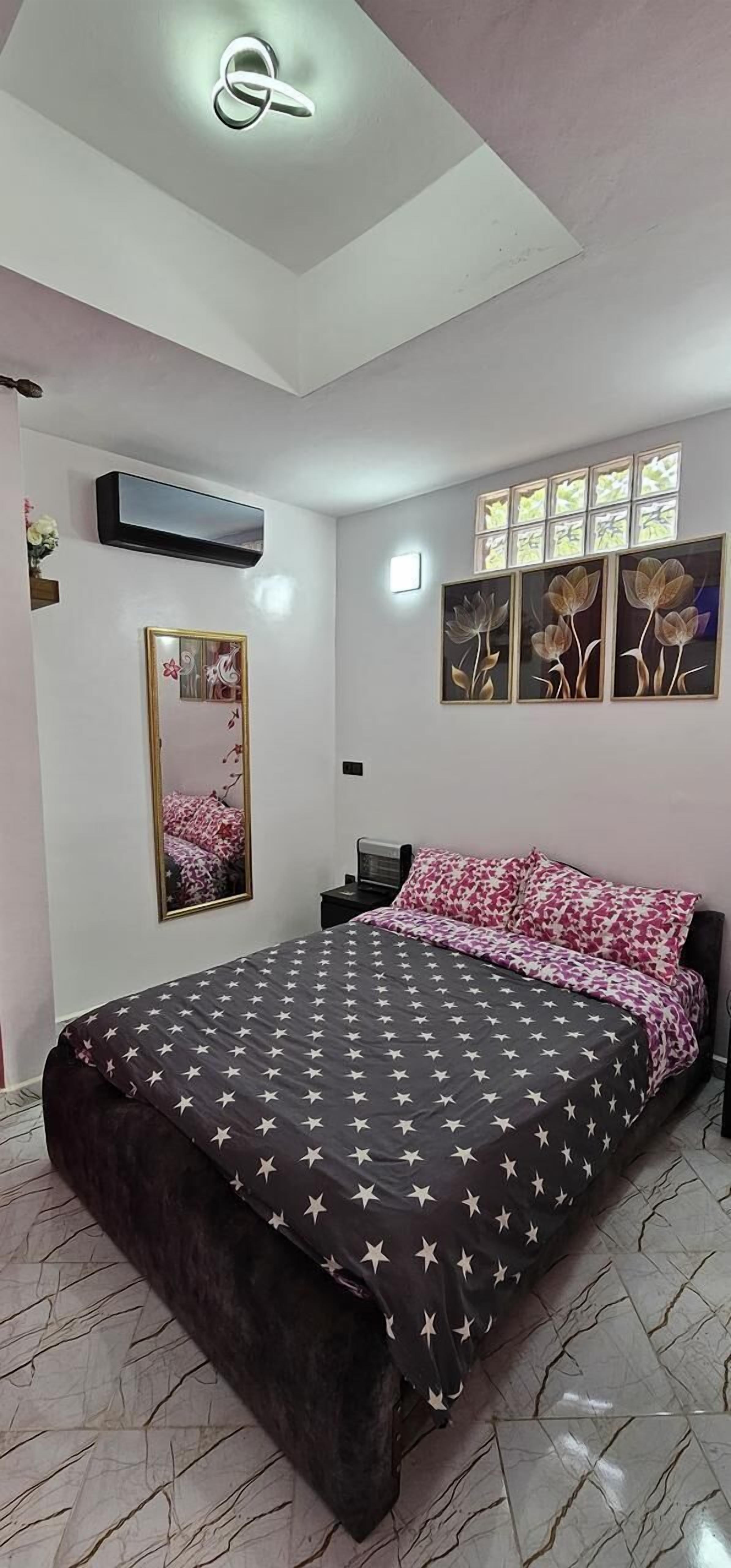 A bedroom with a bed, a mirror, a window with flower paintings, and an air conditioning unit.