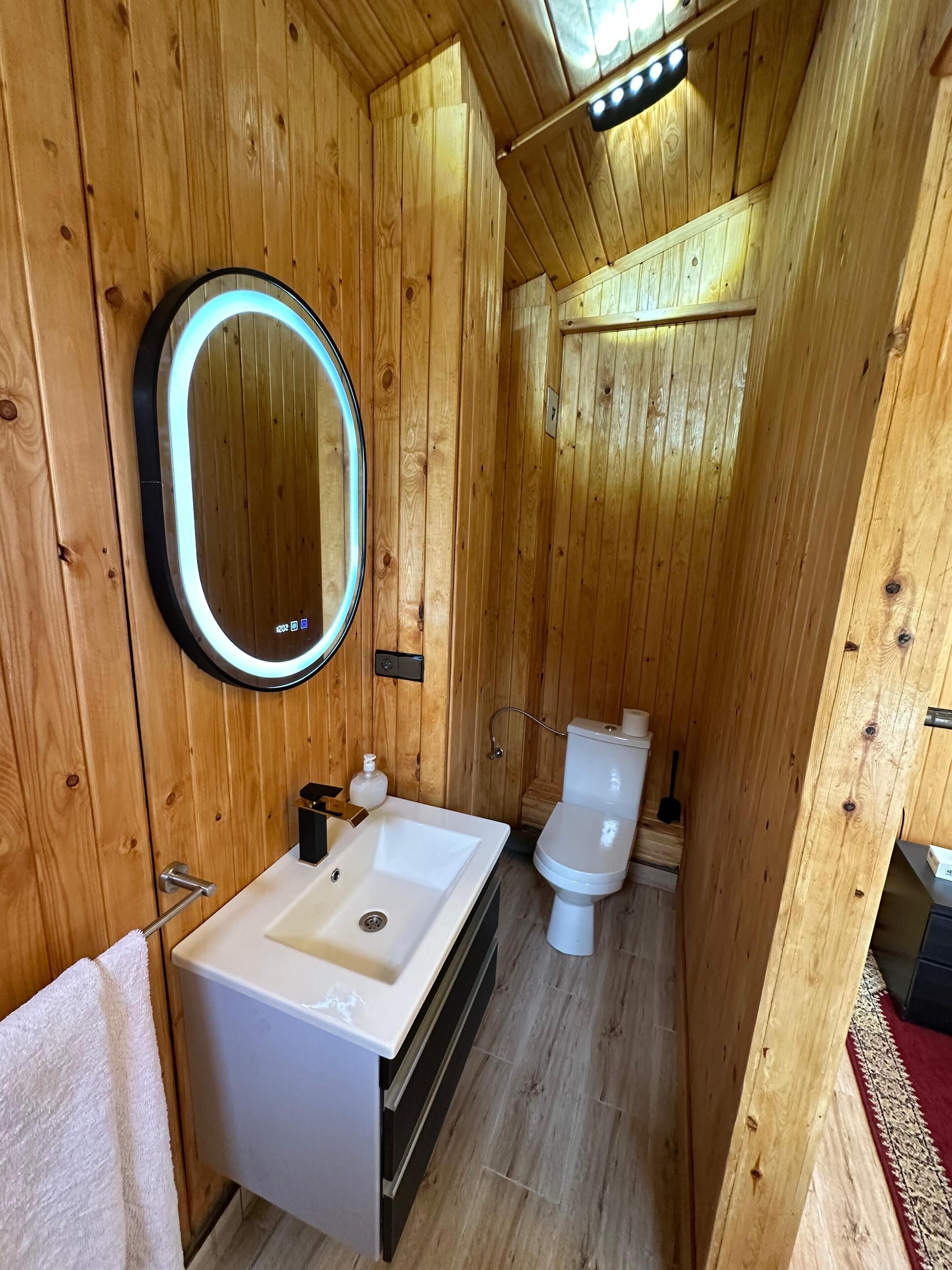 Une petite salle de bain en bois, dotée d’un lavabo blanc, de toilettes et d’un miroir rond avec éclairage à DEL.