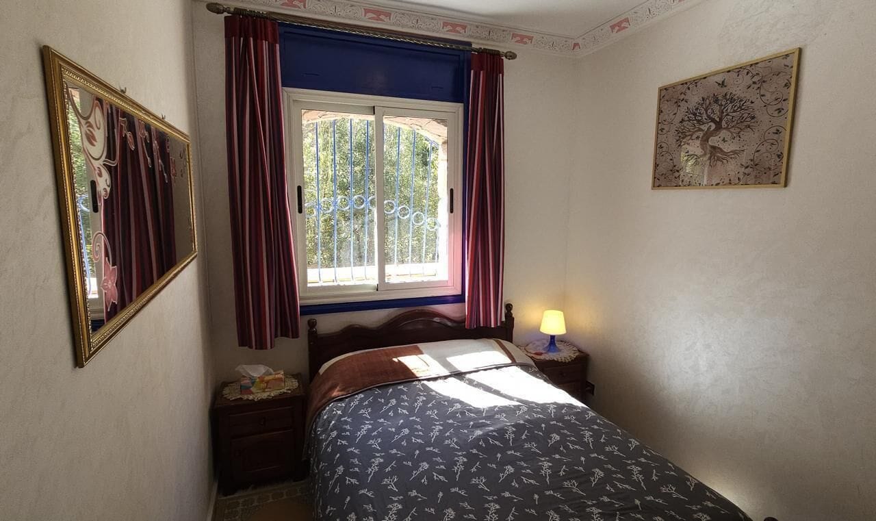 A bedroom with a bed, a nightstand, a lamp, a window with curtains, and a wall-mounted picture.